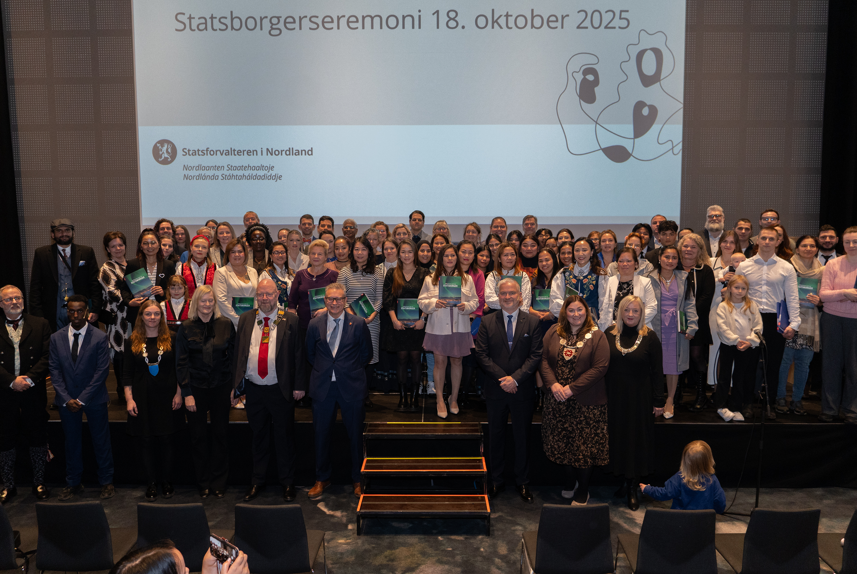 Under seremonien samles deltakere og talere på scenen til gruppebilde. Her fra seremonien i Bodø i 2025. Foto: Emil Harjo Indsetviken / Statsforvalteren i Nordland. En stor gruppe pent kledde mennesker står oppstilt på en scene.