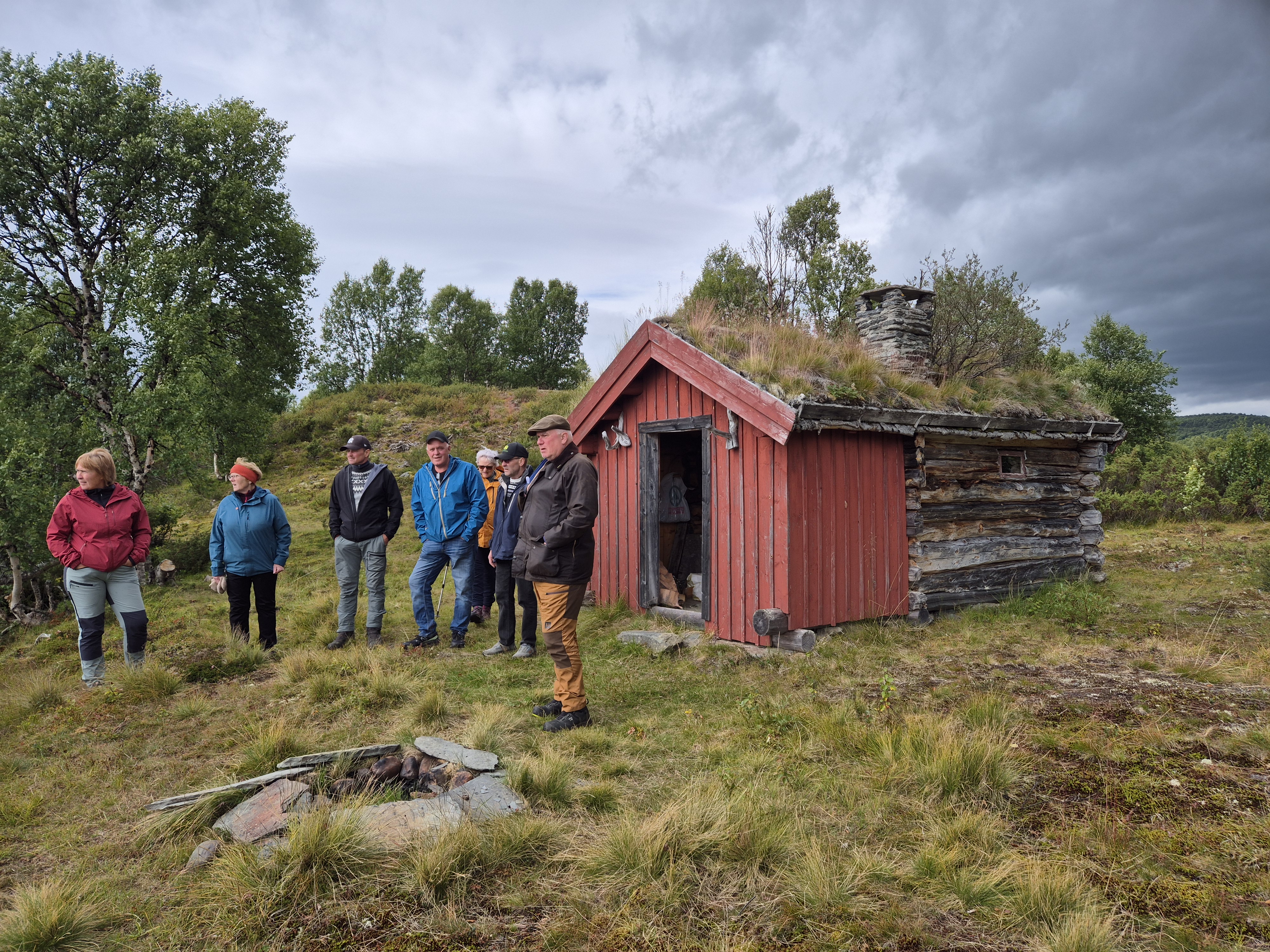 Sju personer som står på naturlig grasmark med bjørker i bakgrunnen, ved siden av et lite tømmerhus med påbygg med rødmalt kledning og gråsteinspipe.