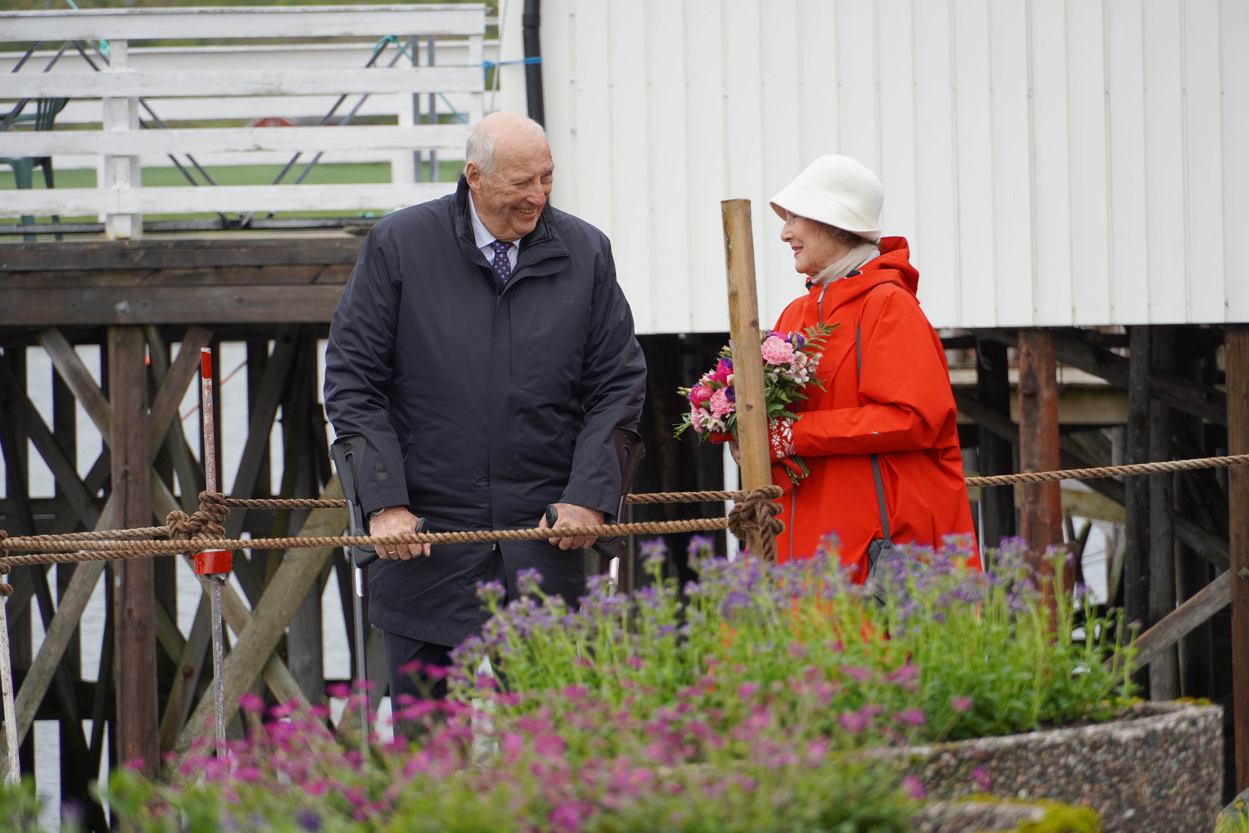 En eldre mann med mørk frakk og en eldre kvinne med rød frakk står foran et hvitt sjøhus.
