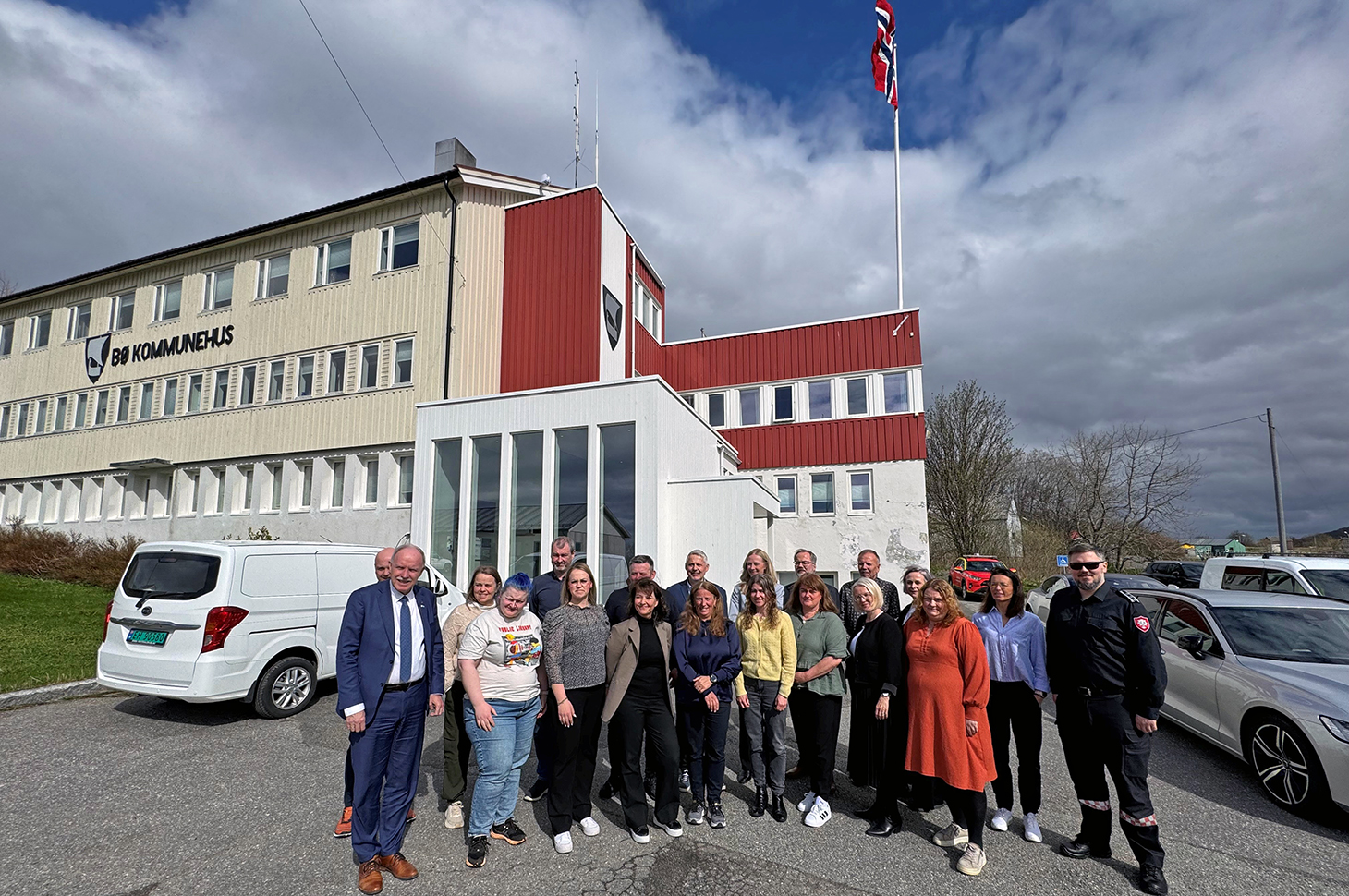 Møtedeltakerne samlet utenfor kommunehuset i Bø. Flagget var heist for anledningen. Foto: Bø kommune. En gruppe mennesker står oppstilt foran en beige, hvit og rød kontorbygning. Noen biler står parkert i området.