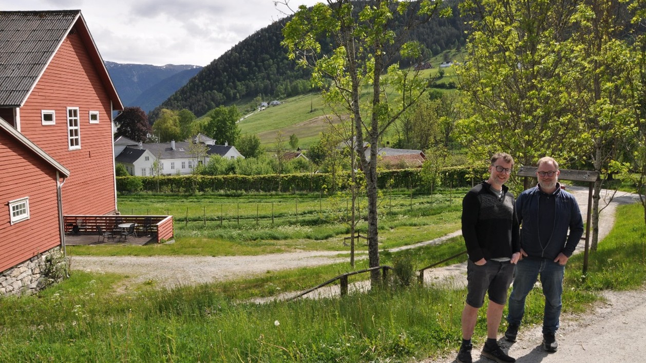 To personer i forgrunnen og gårdslandskap i bakgrunnen med hus til venstre