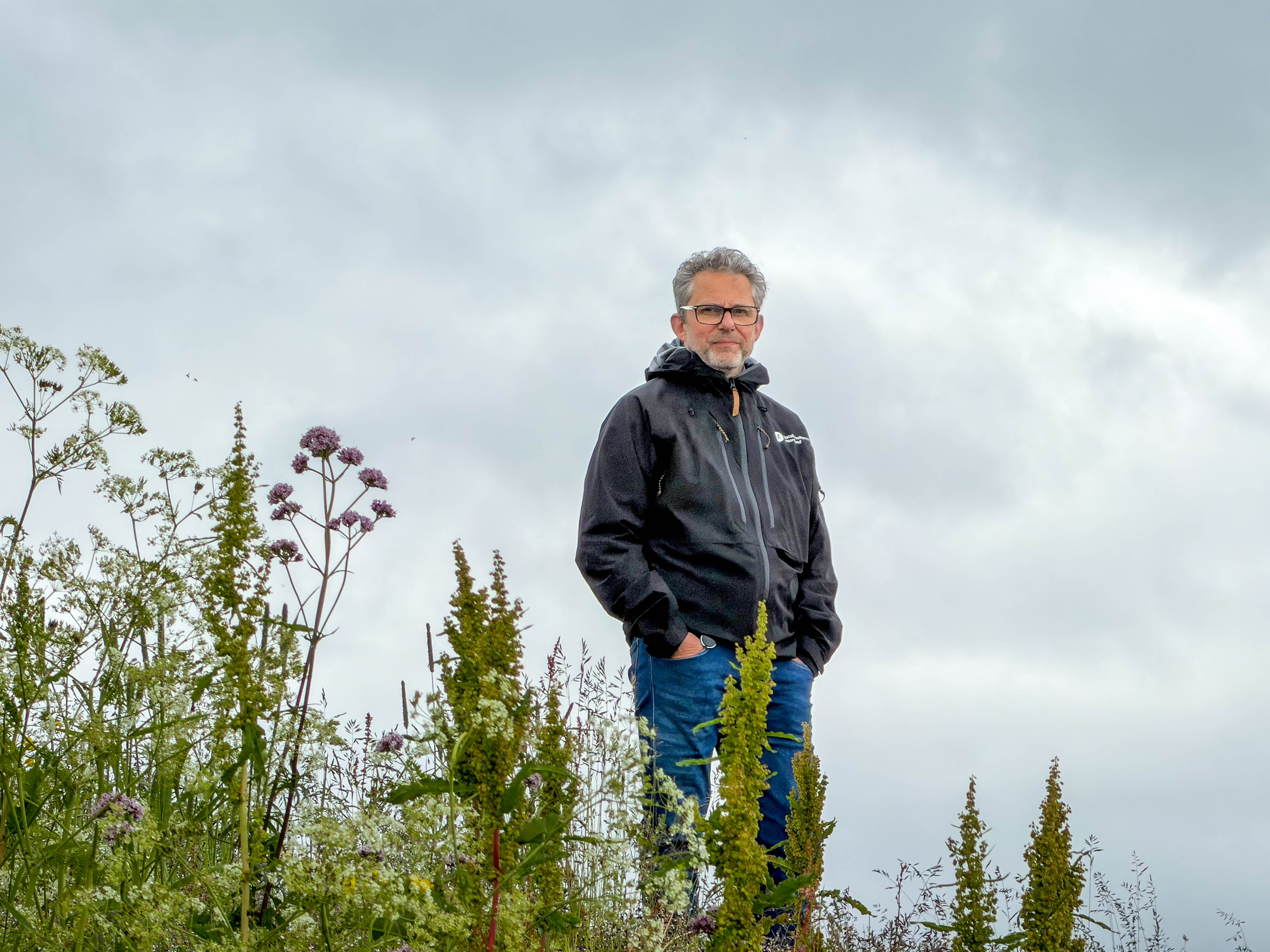 En mann med sort jakke står med hendene i lomma med høyt gress i forgrunnen. 