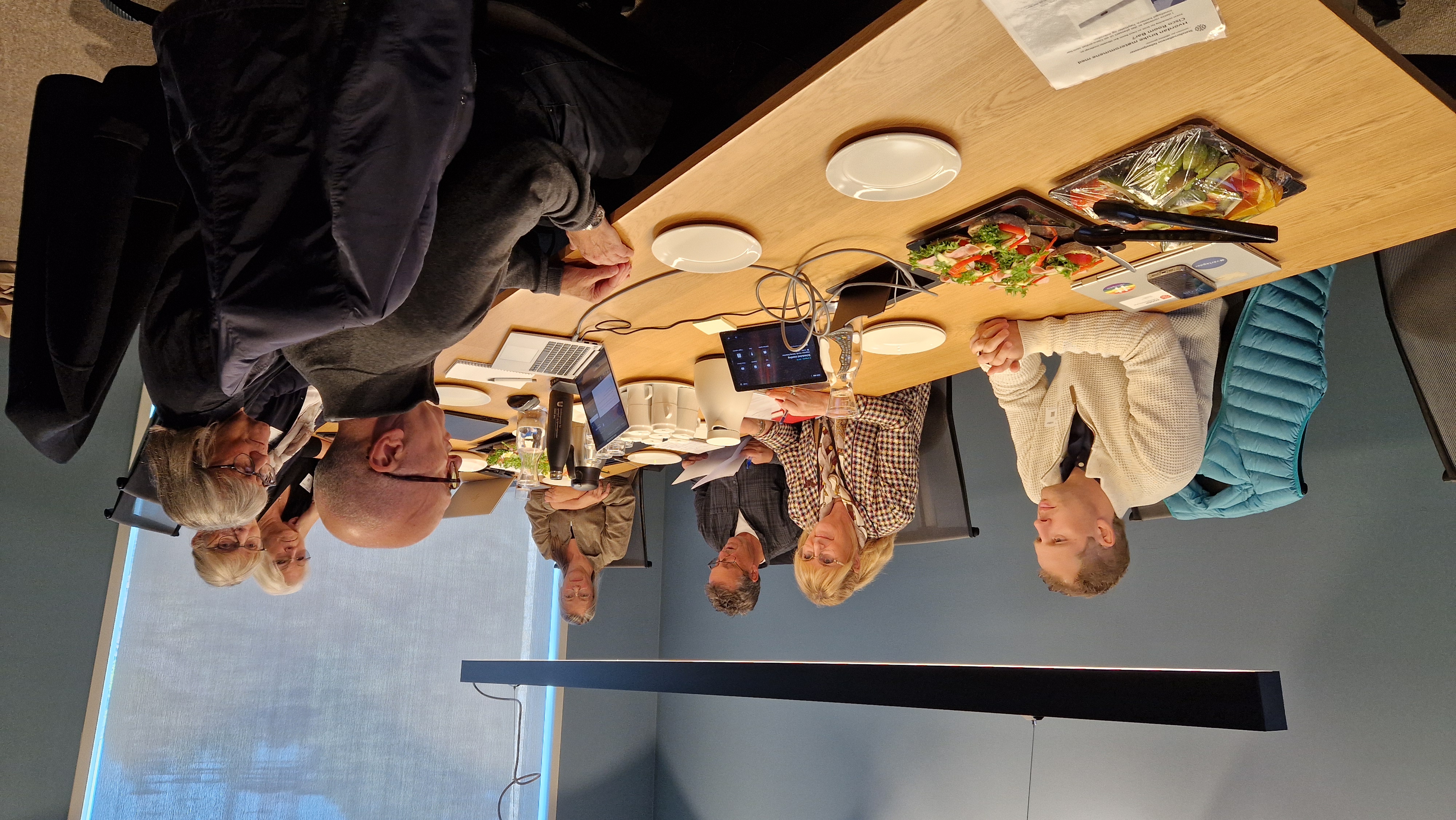 Gruppe mennesker rundt møtebord, med PC-er, kaffekanne og papirer