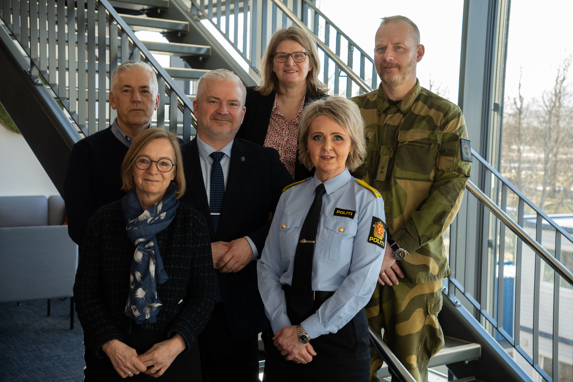 Seks personer står oppstilt ved en trapp med vinduer i bakgrunnen.