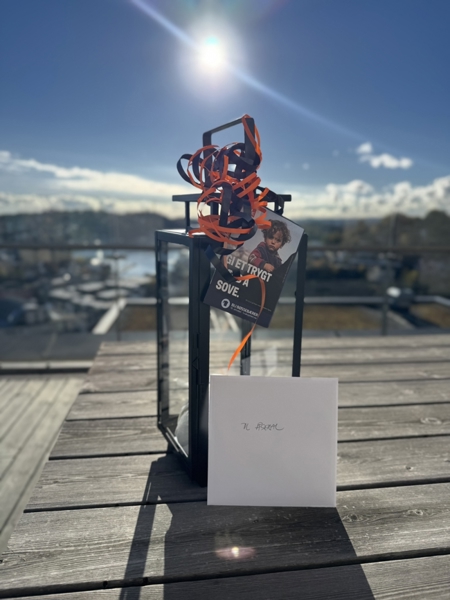 Ei svart lykt pynta med oransje band står på eit trebord utandørs. Framfor lykta står eit kort merka «Til Åseral». Ei brosjyre frå TV-aksjonen er festa til lykta, og i bakgrunnen er det utsikt mot sjø og busetnad i sollys.
