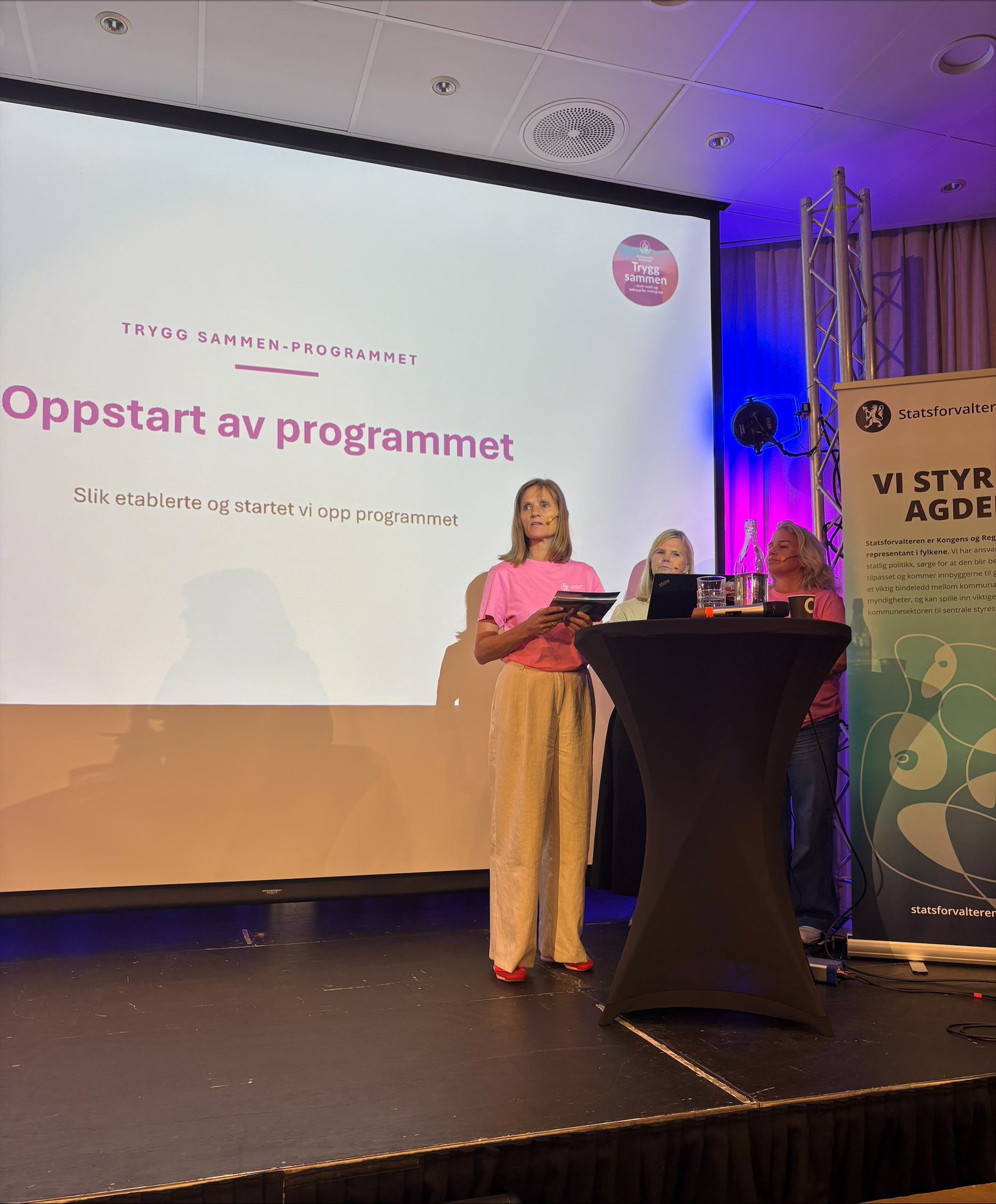Tre representanter fra Kristiansand kommune står på scenen under et seminar og presenterer Trygg Sammen-programmet. På storskjermen bak vises et lysbilde med tittelen «Oppstart av programmet» og teksten «Slik etablerte og startet vi opp programmet». Innlegget handler om kommunens tverrfaglige arbeid for å forebygge vold og seksuelle overgrep mot barn og unge.