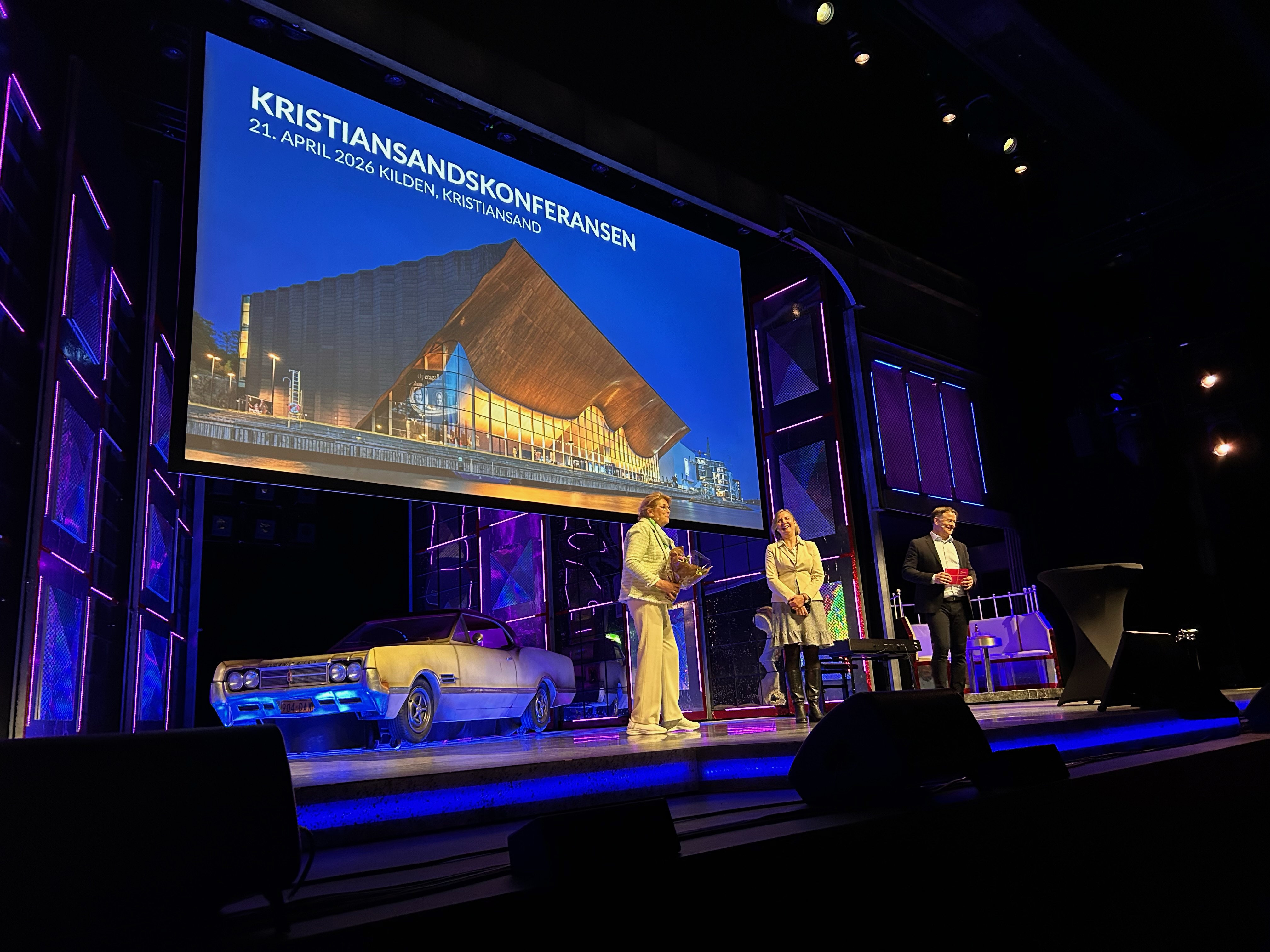 Bilde av smilende mennesker på scene, en av dem med blomster i hendene.