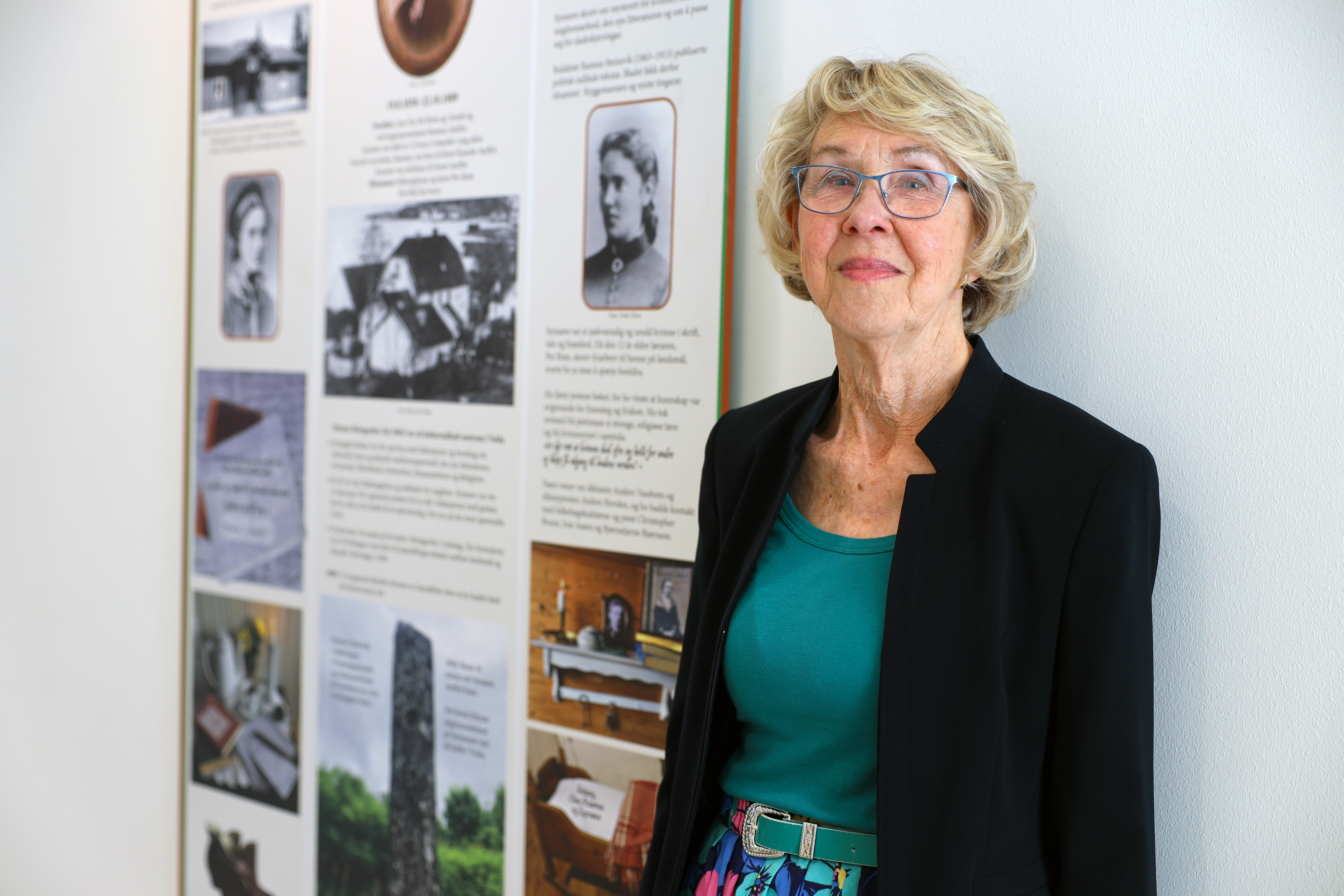 Reidun Aambø har laga ein lærerik poster om namnemora til Synnøve Riste-huset ved Høgskulen i Volda. Foto: Foto: HVO / Steinarr Sommerset. Reidun Aambø står framfor ein poster som ho har laga om Synnøve Riste.