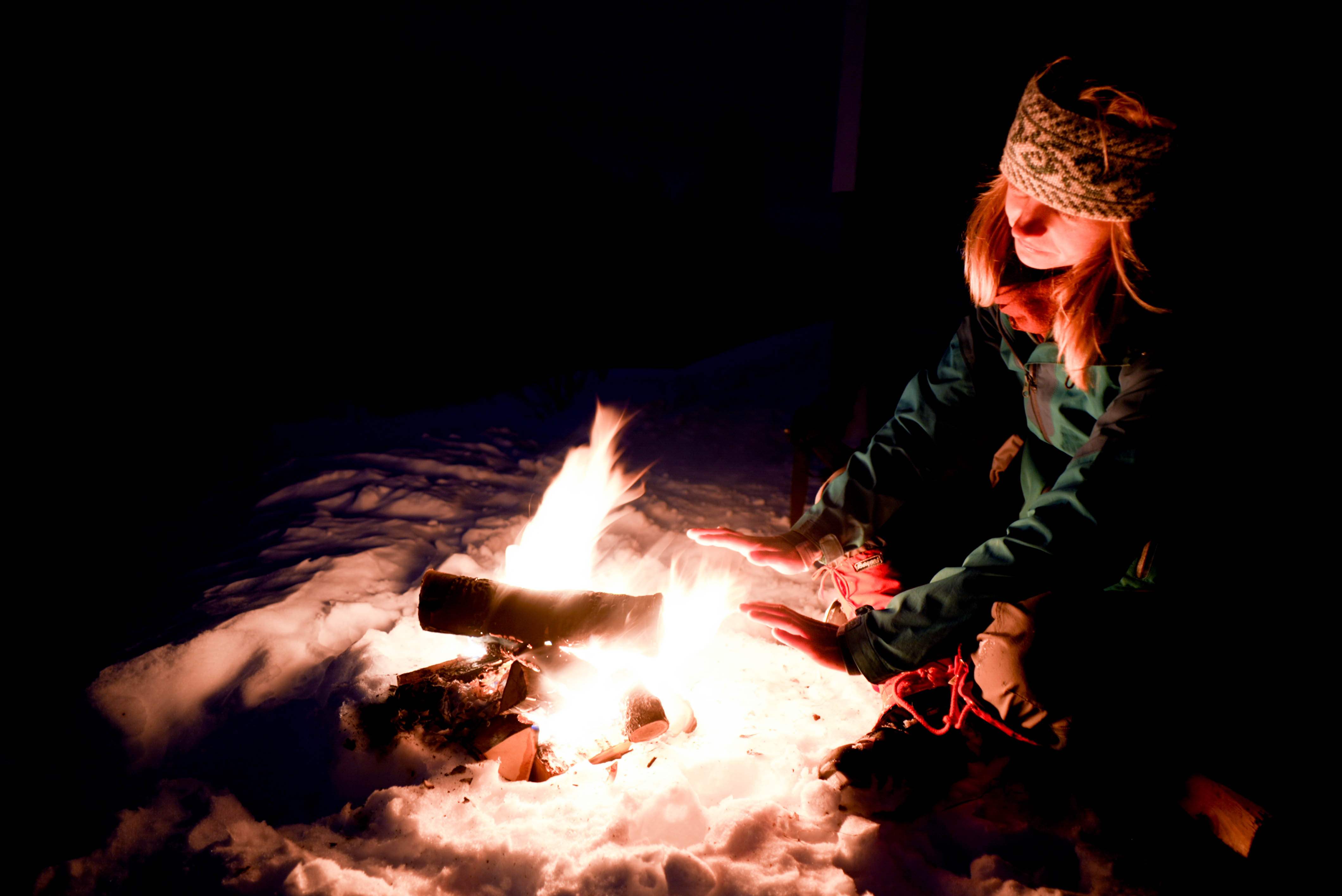 En person sitter på snø foran et bål i mørket og varmer hendene.