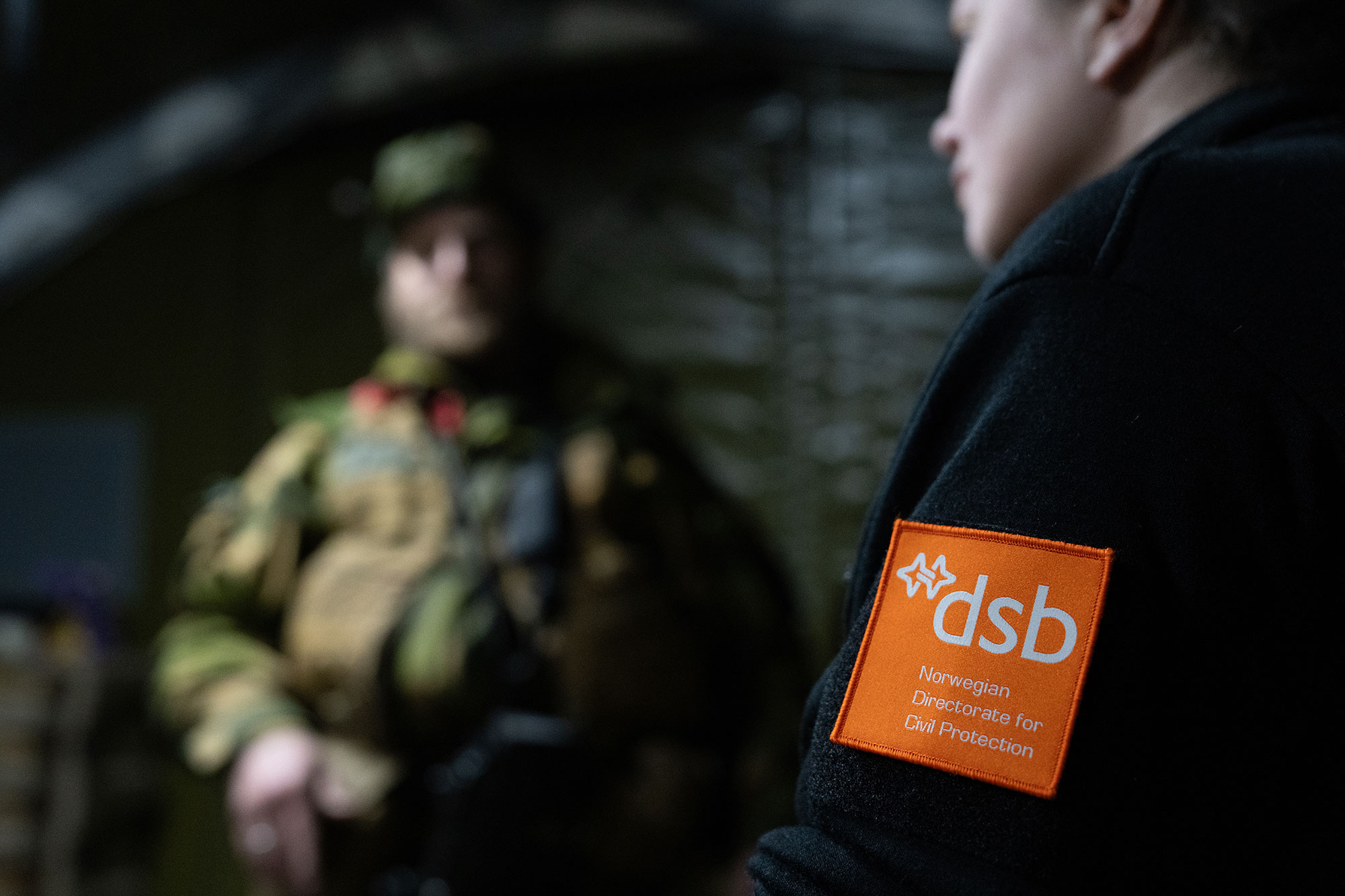 Nærbilde av et emblem med bokstavene dsb på oransje bakgrunn på et jakkeerme. I bakgrunnen står person i kamuflasjeantrekk.