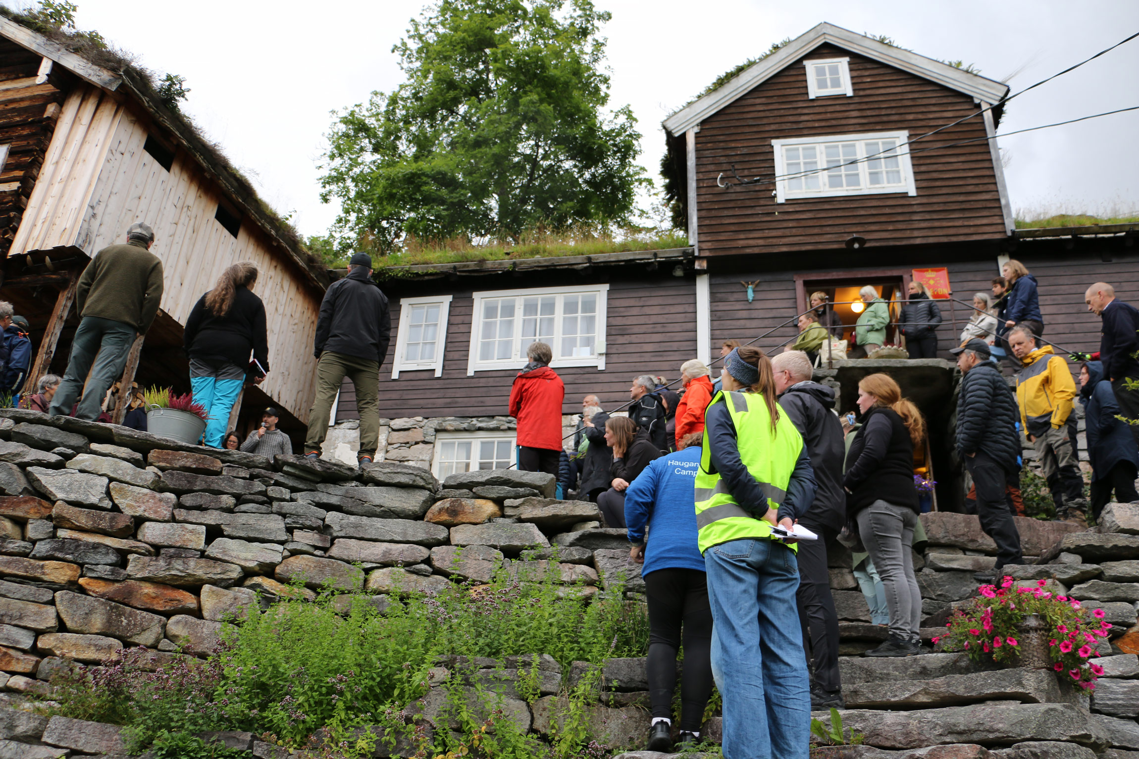Ei gruppe personar står framfor eit gardshus. En mann med mikrofon snakkar til gruppa.