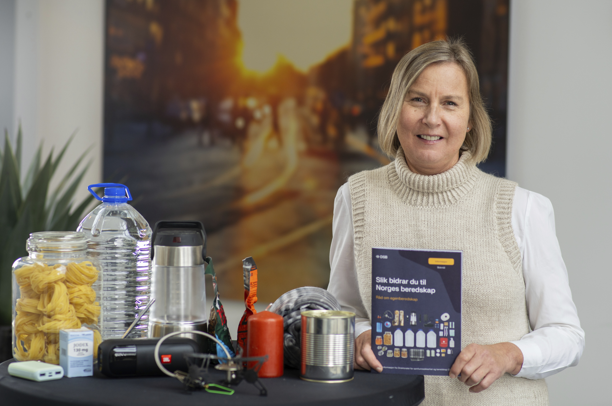 Bilde av Elisabeth Aarsæther som holder brosjyre med tittelen "Slik bidrar du til Norges beredskap". Til venstre et bord med beredskapsutstyr som vann, powerbank mv.