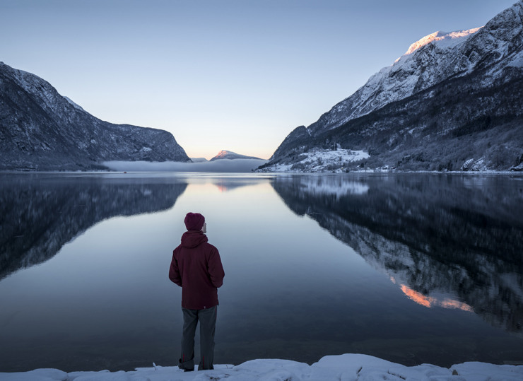 Ein person med raud lue står med ryggen til fotografen og ser ut over eit fjordlandskap med fjell på begge sider av fjorden