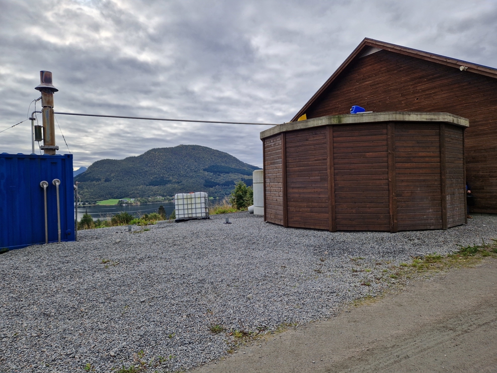 Biogasskum inntil hus, begge kledd i brunbeisa treverk. Utsikt mot fjell i bakgrunnen.
