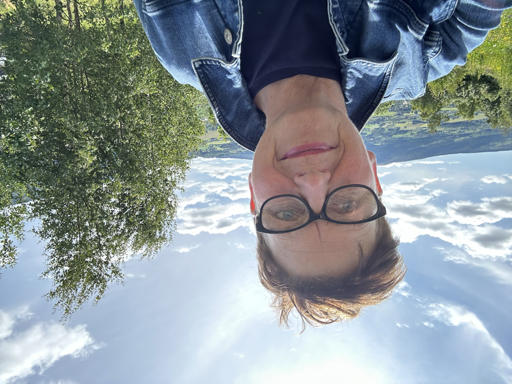 Kvinne kledd i dongeri jakke. Bjørketre og blå himmel bak.